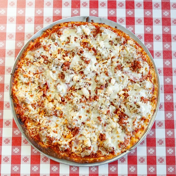 A whole pizza with melted cheese, chicken, and sausage on a red and white checkered tablecloth.