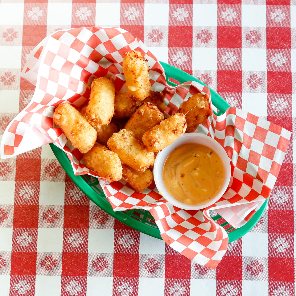 Golden-brown fried cheese curds in a green basket with red and white checkered paper, served with dipping sauce.