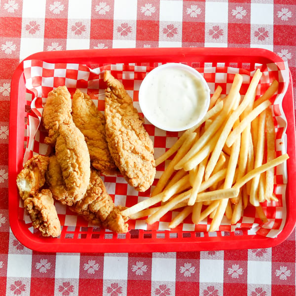 Crispy chicken tenders, golden fries, and a cup of ranch dressing in a red basket on a checkered tablecloth.