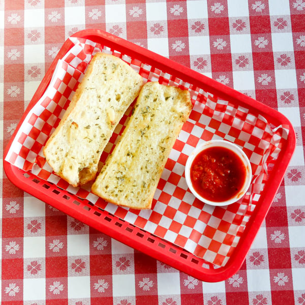 Two slices of garlic bread with a side of marinara sauce in a red basket on a checkered tablecloth.
