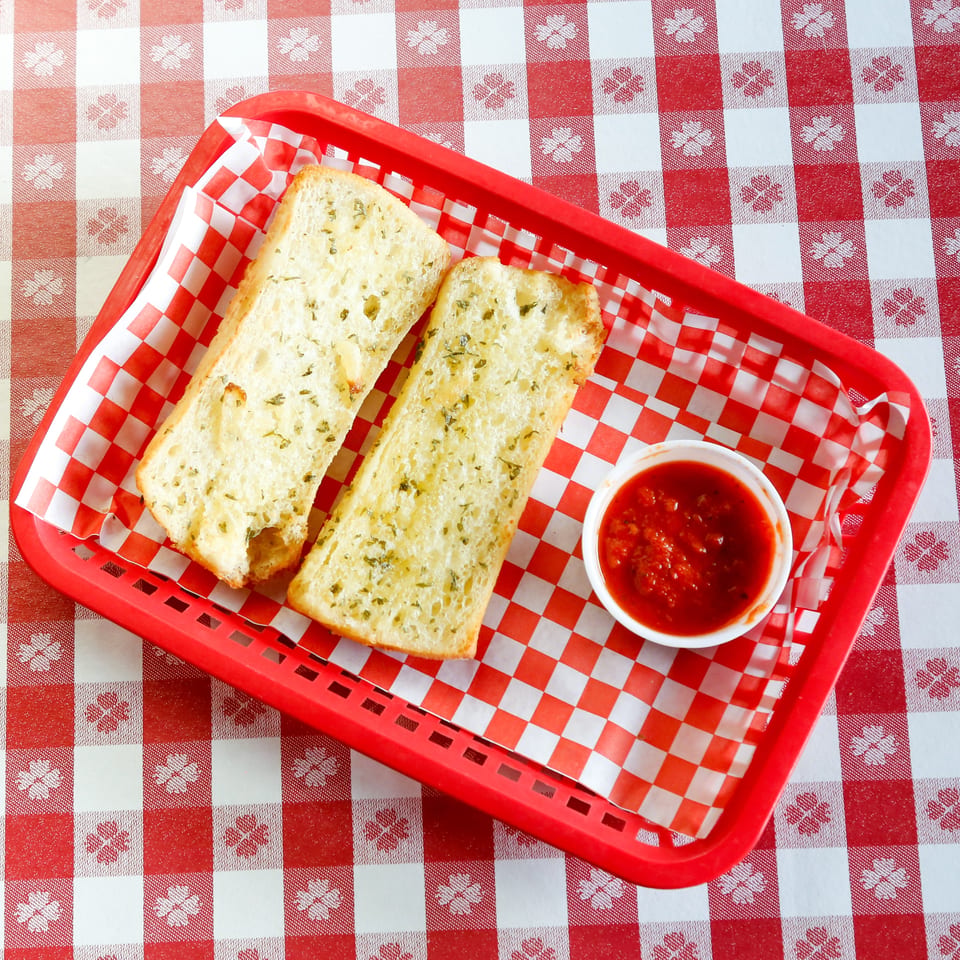 Two slices of garlic bread with a side of marinara sauce in a red basket on a checkered tablecloth.
