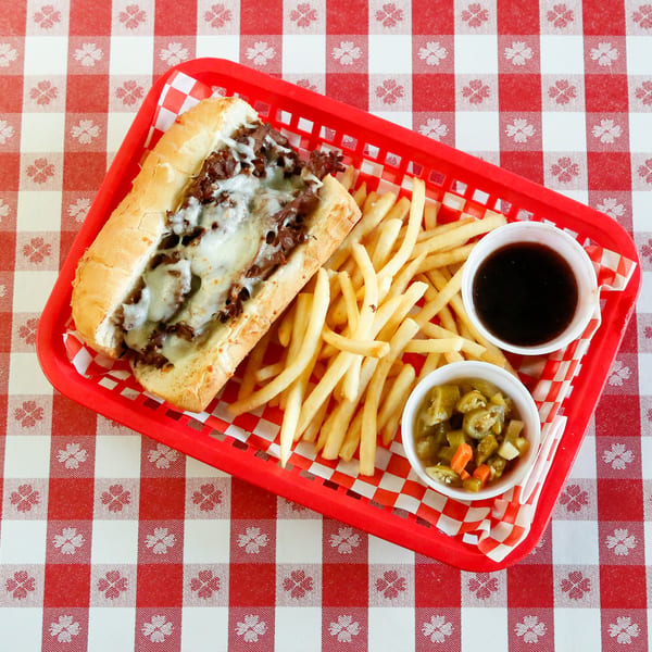 Cheesesteak sandwich with fries, au jus, and pickled vegetables in a red basket on a checkered tablecloth.